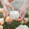 Fresh Face Cream in a jar held gently in hands, surrounded by soft flowers. Anti-aging and plumping moisturizer.