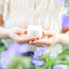 Fresh Face Cream Tallow Moisturizer in a jar, held among soft purple flowers, ideal for anti-aging skincare.